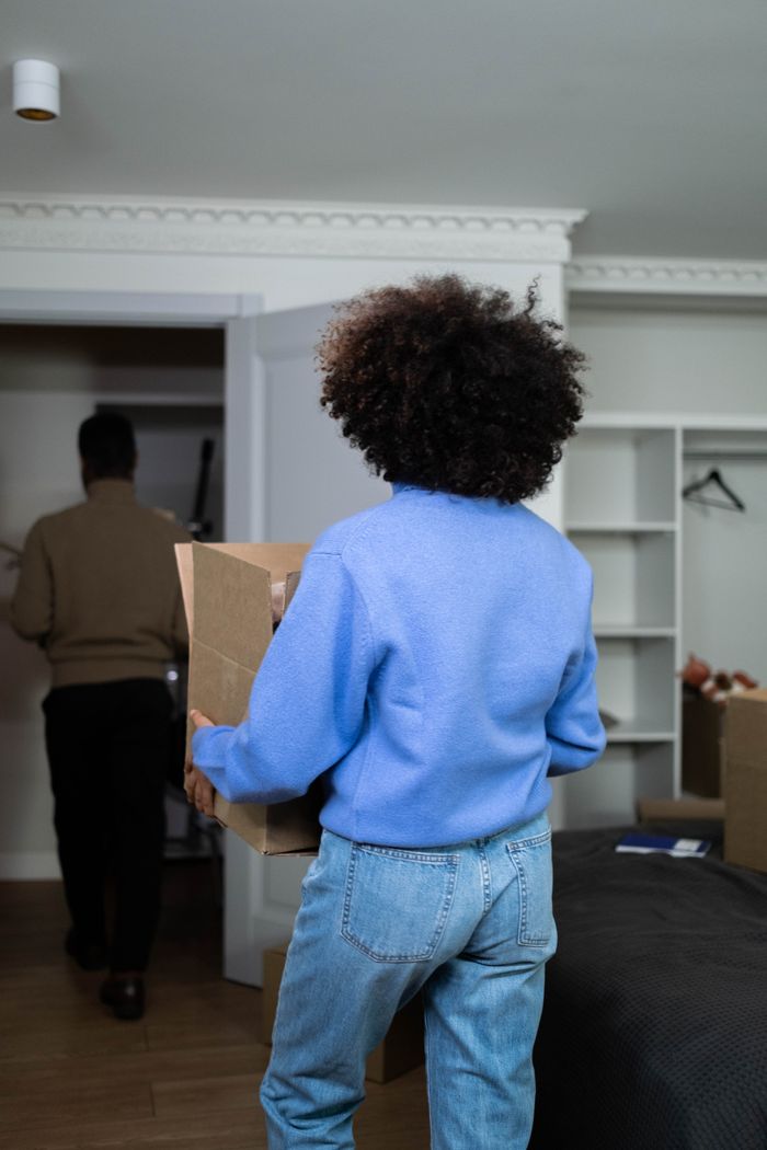 Vue arrière d'une femme portant une boîte à la maison lors d'une journée d'emménagement, mettant en avant le thème du déménagement et du transfert.